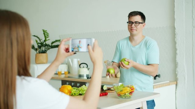 Student handsome man in glasses is juggling apples in kitchen when girlfriend taking pictures with smartphone camera in kitchen in apartment. Youth and fun concept.