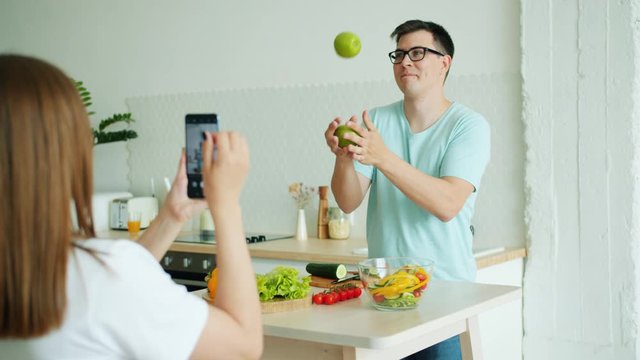 Slow motion of handsome guy juggling apples then posing for smartphone camera while woman taking photos in kitchen in modern apartment. Youth and lifestyle concept.