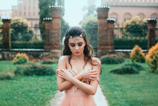 Attractive brunette girl with a cute angel face standing in the rain under the open sky. A young princess in a pink dress is sad, frozen, wet, hugs herself on the shoulders. Art photography