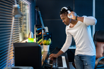 Young serious casual mixed-race man with headphones standing by computer screen