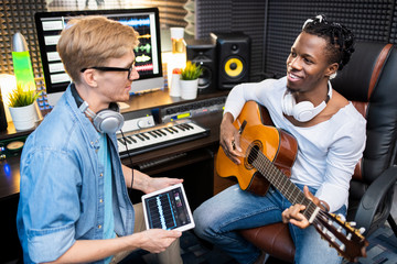 Young man with digital tablet talking to happy colleague with guitar