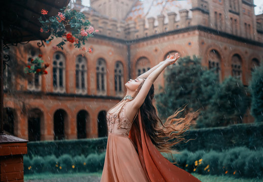 Lady with a pretty face, stands under the rainy sky and reaches out to meet the drops. Young magician in a orange dress with a cape near the castle. Dance with the wind, long hair flying, fluttering.