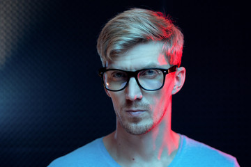 Young serious man with blond hair standing by wall in dark studio in isolation