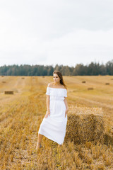 Fototapeta na wymiar Beautiful girl in a long white dress and barefoot standing at setga of straw on a yellow field in summer