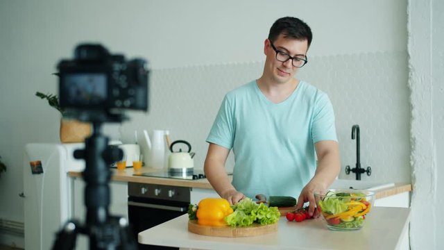 Young handsome man vlogger is recording video about organic food cooking vegetable salad in kitchen at home using camera. People and nutrition concept.