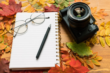 Photocamera, blank page of copybook with eyeglasses and pen and autumn leaves