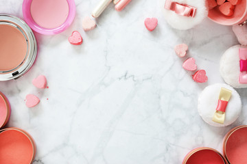 Flat lay on marble background with cute pink beauty products like cute powder puffs with ribbons, blush pots, makeup brushes and pressed powder in heart shape arranged around copy space