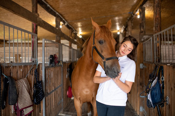 Young active woman in white shirt standing inside stable by purebred racehorse