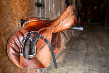 Brown shabby leather saddle with black bridles hanging on steel bar