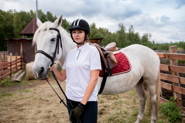 Young sportswoman in equestrian outfit standing by white purebred racehorse