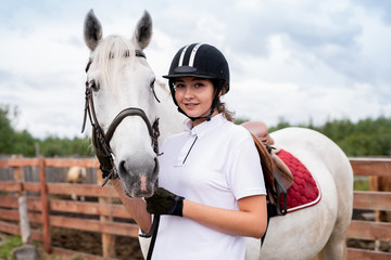 Young smiling woman in equestrian outfit standing close to white racehorse