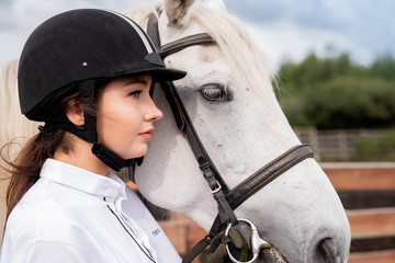 Young calm female in white shirt and equestrian helmet standing by racehorse