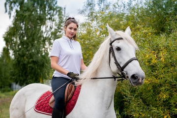 Pretty girl looking at you while sitting on back of white purebred racehorse