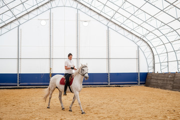 Young active woman sitting on back of horse while moving down sandy arena