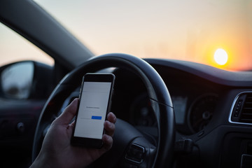 Close-up of male hand hold smartphone inside car against steering wheel at sunrise. No network...