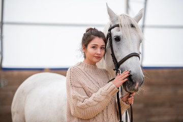 Calm young casual woman in knitted sweater standing by white purebred horse