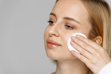 Young blonde young woman cleaning (removing makeup) her face with cotton pad, isolated on gray background, copy space. Healthy skincare, facial treatment