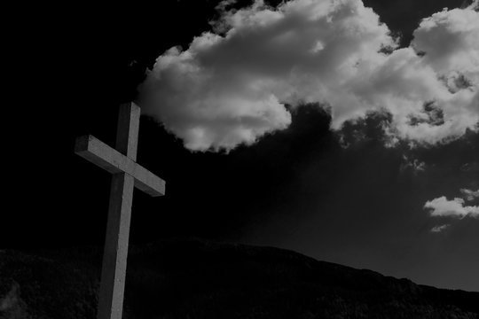 Close up cement cross(Jesus Christ crucifix) with mountain ranges background, thick white clouds on the top of the rood crucifix. Black and white image.