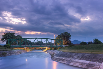 Lighten, iconic, metal railroad bridge in Pirot, Serbia,  over silky water river Nisava and dramatic sunset sky