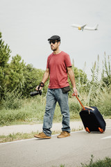 Traveler walking around with a camera in his hand and a backpack. There is a plane in the sky