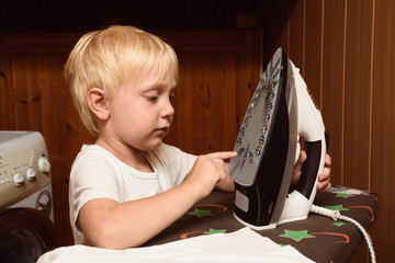 Fototapeta na wymiar Little blond boy touches electric iron with his finger. Home safety