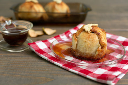 Freshly Baked Homemade Apple Dumplings in Apple Shaped Plate with Red and White Checked Napkin on Wooden Table; Apple Shaped Bowl of Brown Sugar Cinnamon Syrup in Background