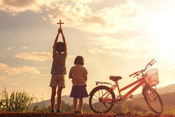 Children standing on mountain praying and holding christian cross for worshipping God at sunset...