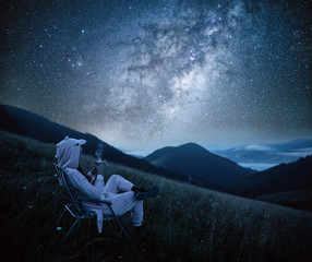 Young woman drinking coffee at mountains on the background of the milky way. Woman looking at beautiful milky way. Starry sky with hills at summer