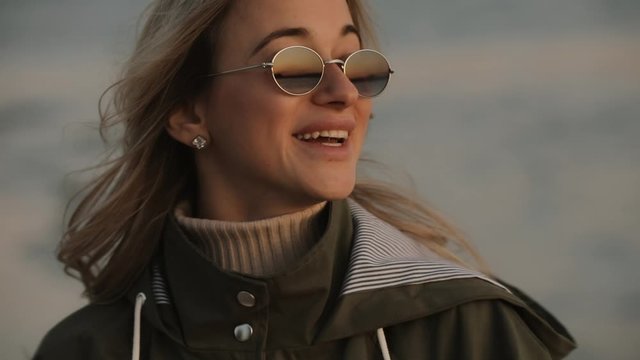 Stylish happy woman in sunglasses laughing standing at the river at fresh autumn evening
