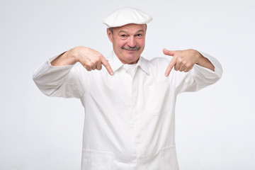 Smiling senior cook in white uniform smiling and pointing down trying to pay attention to important information.