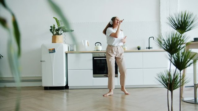 Slow motion of cute girl in wireless headphones singing in spoon in kitchen dancing having fun alone. Modern technology, lifestyle and people concept.