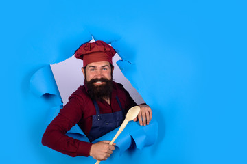 Handsome chef cook with wooden spoon looking through paper. Chief man in cook uniform holds wooden kitchen cooking tools. Bearded man chef preparing to cook food. Bearded cook with kitchen utensils.