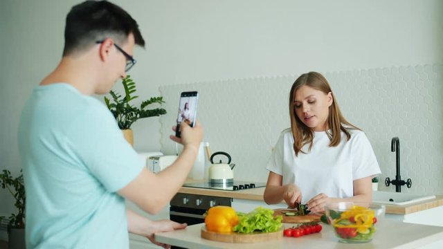 Slow motion of young woman cooking salad having fun eating cucumber while man taking photo with smartphone. People, lifestyle and healthy nutrition concept.