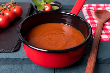 Molho de tomete na panela vermelha com fundo preto em cima da madeira preto e verde petróleo com colher de madeira, tomate cereja na ardósia negra e folhas de majericão