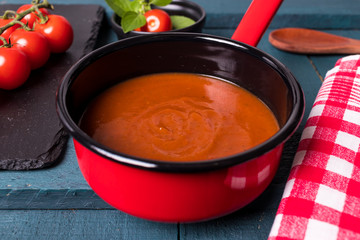 Molho de tomete na panela vermelha com fundo preto em cima da madeira preto e verde petróleo com colher de madeira, tomate cereja na ardósia negra e folhas de majericão