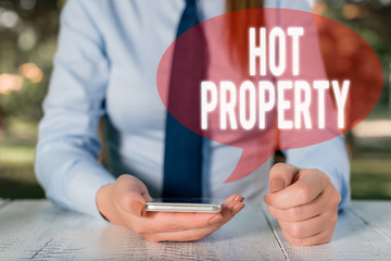 Writing note showing Hot Property. Business concept for Something which is sought after or is Heavily Deanalysisded Female business person sitting by table and holding mobile phone