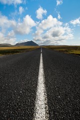 Low angle shot of an icelandic road extending to then horizon, with distant mountains and under a blue sky with puffy clouds