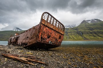 Icelandic landscape with a rusty wrecked ship stranded on a fiord in foreground and distant snow-covered mountains in the background, under a dark grey sky