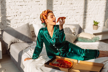 young cheerful woman in silk nightwear eating tasty pizza in bed, unhealthy eating