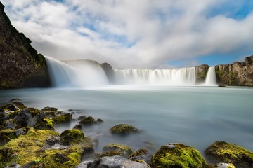 Icelandic landscape portraying the great waterfall of Godafoss, with musk-covered rocks in foreground and under a blue sky with puffy clouds