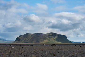 Icelandic landscape with a flat desert and a single mountain, under a blue sky with puffy clouds
