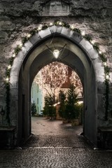 Naklejka premium An ancient stone gate with a marble pointed arch decorated with christmas lights; beyond the gate there are two trees adorned with lights
