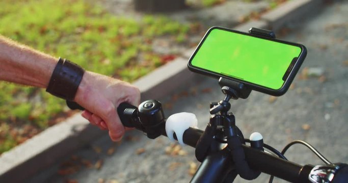 Close-up young man riding bicycle using mounted smartphone with mock-up green display outdoors on sunny evening.