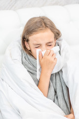 Sick girl wrapped in a white blanket sitting on the bed and blowing her nose in tissue