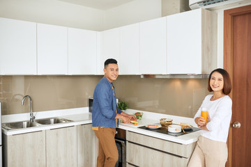 Young couple having breakfast, woman holding orange juice while man cooking food kitchen modern apartment interior
