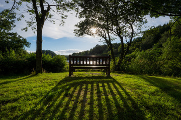 In a meadow overlooking a scottish lake at sunset, a wooden bench is casting an elongated shadow