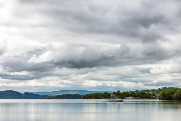 A scottish lake surrounded by trees and vegetation, with a motorboat moored in the middle and distant mountains under a cloudy sky