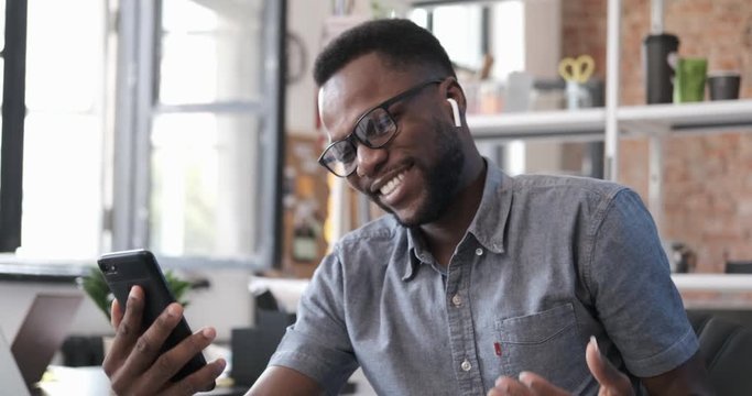 Happy african american man smiling and emotionally talking with his friend on smartphone. Man having video chat at office. Enjoying communicating. People and modern technology. Indoors 4k close up