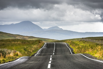 A narrow scottish road is extending to distant mountains covered by haze