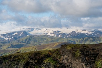 Icelandic landscape with a green mountain ridge in the foreground, and distants snow-capped mountains in the distance, under a blue sky with grey clouds